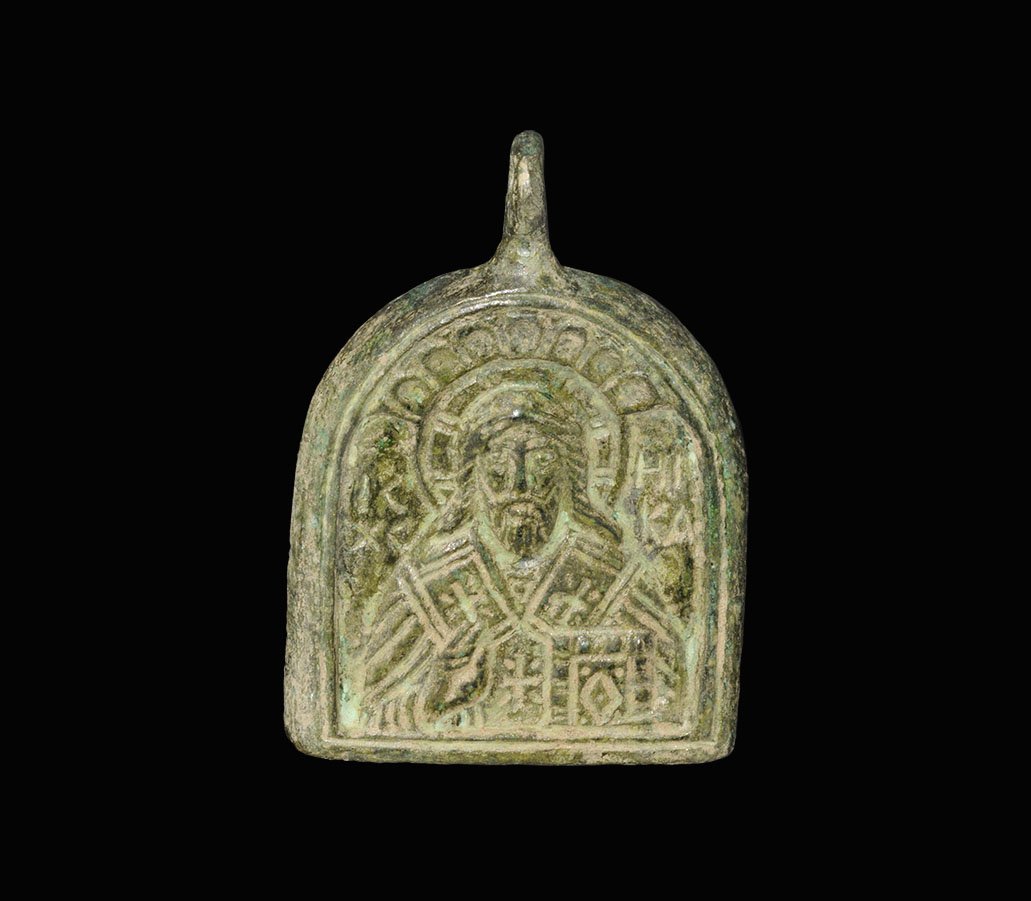 Byzantine Style Bronze Pendant: An undated archaistic cast D-shaped pendant plaque with high-relief figure of Christ with right hand raised in blessing, left hand holding a book, nimbus to the rear, incuse text 'IC / HI / XC / KA' f