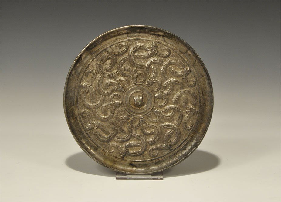Chinese Style Bronze Serpent Mirror: An undated archaistic discoid mirror with one polished face; to the reverse a central pierced lug surrounded by a band of sixteen intersecting serpents. 746 grams, 17.5 cm (7"). London private collect