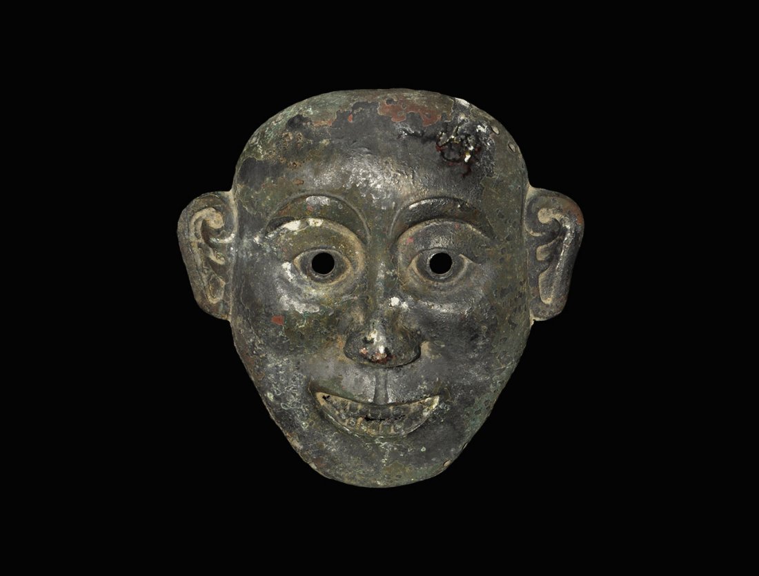 Chinese Style Bronze Face Mask: An undated archaistic cast face mask in Han Dynasty style, with round eyes, triangular nose and grinning mouth; pierced at the pupils and to the edges four pairs of lace-holes. 674 grams, 19.5 cm (7 3