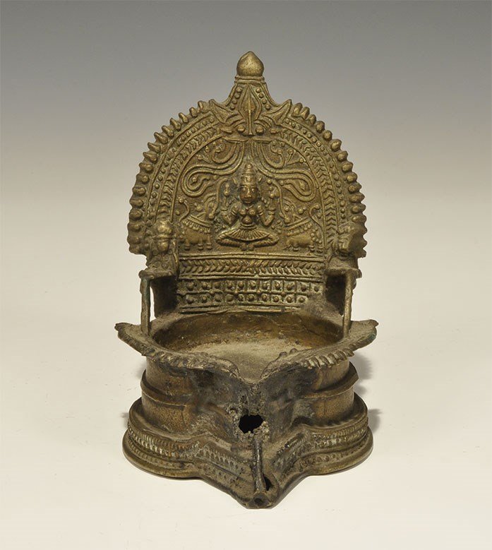 Indian Brass Buddhist Throne Lamp: 19th century AD or earlier. A cast model throne with raised decoration of pellets and chevrons, pedestal bae and D-shaped back with raised goddess figures, bulls and vegetation; the seat a recess with