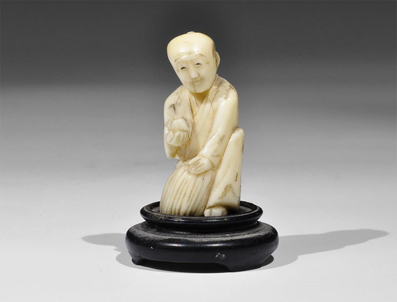 Japanese Antique Ivory Kneeling Figure with Fruit: Late 19th century AD. A carved figurine of a kneeling male in a loose robe with the left hand resting on a sack, a fruit in the right hand; incised geometric motifs to the robe; supplied with a pedest