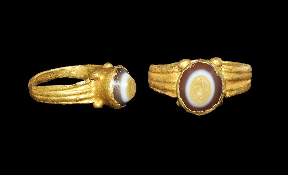 Roman Gold Ring with Trophy on Onyx Intaglio: 1st century AD. A flat-section hoop with expanding ribbed shoulders, funicular bezel with four granules to the base, conical onyx insert with intaglio trophy motif. 2.93 grams, 23 mm overall, 14.85 mm