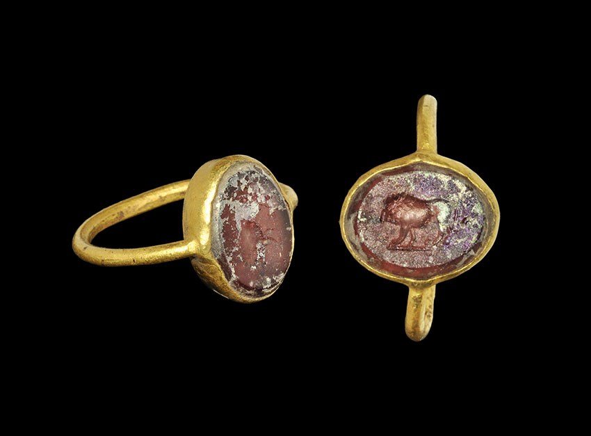 Roman Gold and Mauve Glass Ring with Lion Intaglio: 1st-2nd century AD. A round-section hoop supporting an elliptical bezel with inset mauve iridescent glass panel intaglio motif of an advancing lion. 5.51 grams, 22 mm overall, 20.88 mm internal diamet
