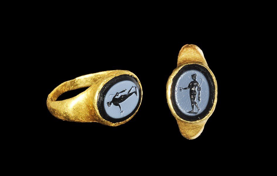Roman Gold Intaglio Ring with Standing Figure: 1st-3rd century AD. A D-section hoop with expanding shoulders, elliptical bezel with inset black stone intaglio depicting a standing helmetted figure with cup in one hand. 5.94 grams, 22 mm overall, 1
