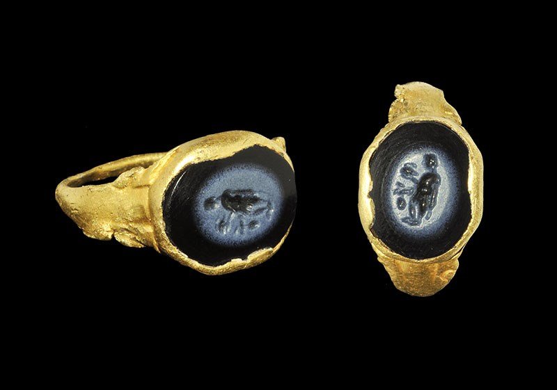 Roman Gold Ring with Goddess Intaglio: 1st-3rd century AD. An elliptical-section hoop with expanding shoulders, scroll detail to the edges and discoid bezel inset with a black intaglio depicting a standing figure of a Roman goddess holding