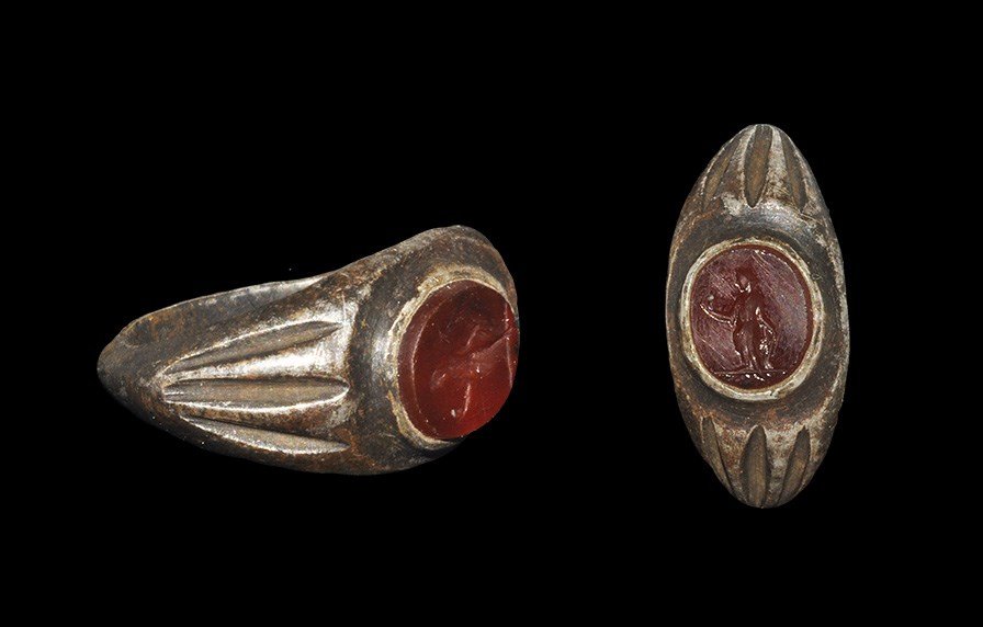 Roman Silver Ring with Carnelian Intaglio of a Goddess: 1st-2nd century AD. A carinated D-section hoop with modelled ribbed shoulders and discoid carnelian to the bezel with intaglio design of a standing female figure, probably Laetitia, with hand extended