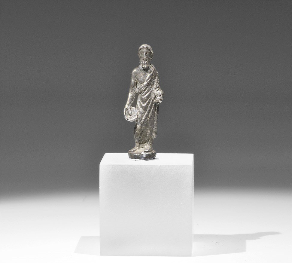 Roman Silver Figurine of a Philosopher: 1st-2nd century AD. A cast figurine of a standing bearded man in a loose robe fastened at the left shoulder, paten in the right hand and palm frond resting on the left forearm; discoid base; on a cust