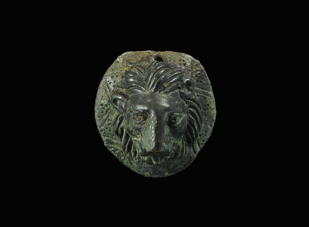 Romano-British Bronze Lion-Head Mount: 2nd-3rd century AD. A cast discoid mount, hollow to the reverse, with a lion's head modelled in the half-round on a field of chevrons and triangular punchmarks; pierced above for suspension. 113 grams