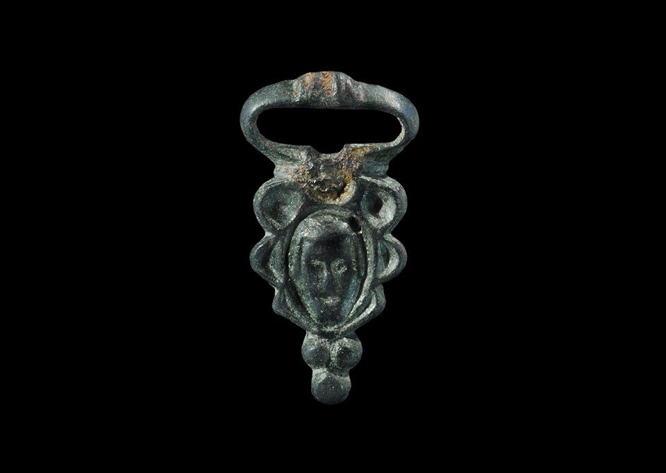 Roman Bronze Buckle with Face: 4th century AD. A cast miniature buckle with triangular plate and elliptical loop; the plate with high-relief face motif with lateral lobes, three pellets below; the loop with couch to the forward edg