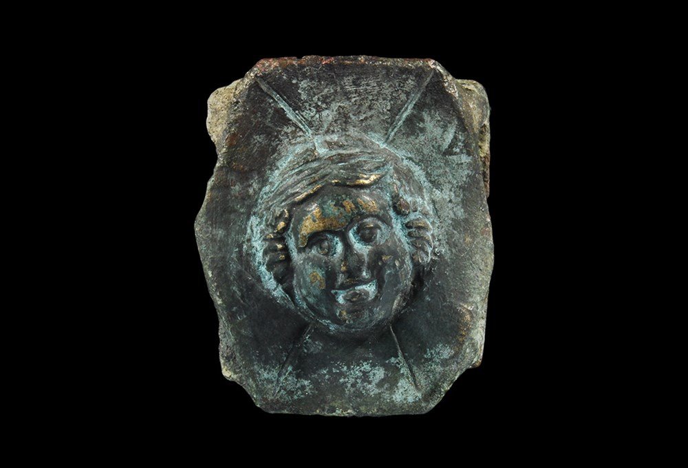Roman Bronze Belt Mount: 1st century AD. A cast hexagonal belt mount with sliding attachment bars to the reverse; the plate with high-relief head of Venus and four radiating incised strokes. 31 grams, 40 mm (1 1/2"). Property