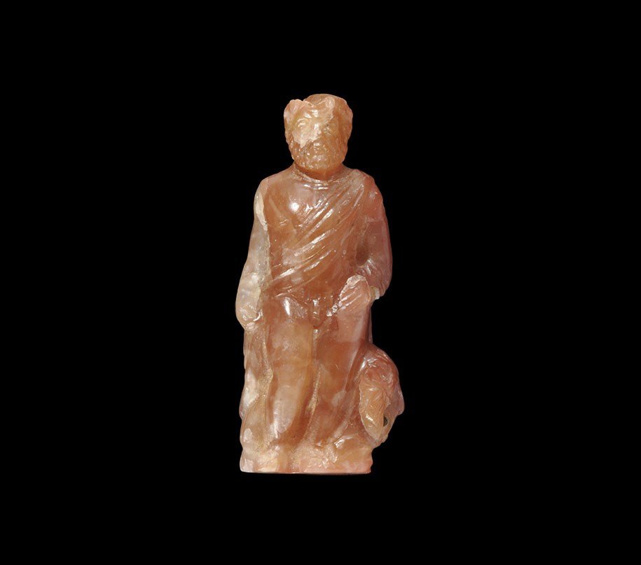Roman Carnelian Hercules Statuette: 1st-2nd century AD. A carved carnelian statuette depicting a nude bearded male standing with a cloak draped over his shoulders, a lion's head at his left foot; the lion's mouth pierced to emphasise th
