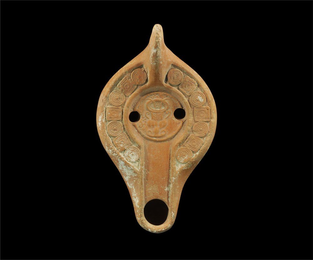 Roman Ceramic Oil Lamp with Amphora: 3rd-5th century AD. A terracotta oil lamp with bulbous body and nozzle, triangular lug handle to the rear; the discus with alternating square and circular panels surrounding a vase with flowers and be