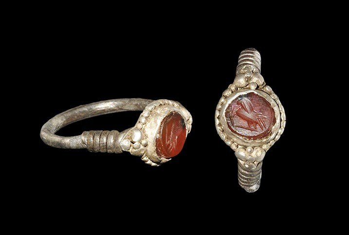 Frankish Silver Ring with Roman Intaglio: 6th century AD. A round-section hoop with coils to the shoulders, discoid bezel with granulated border, inset 1st-2nd century AD hardstone intaglio depicting a standing bird. 3.99 grams, 26 mm overall