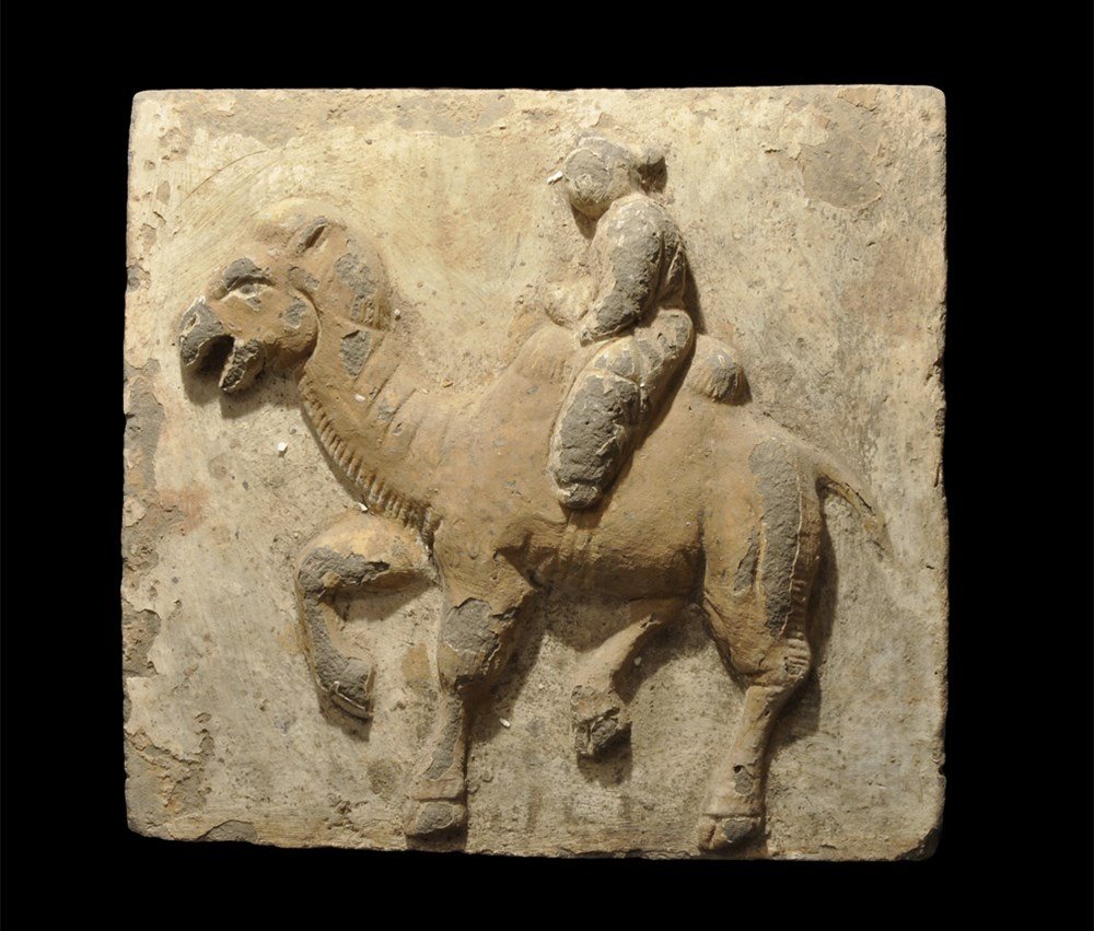 Chinese Polychrome Painted Brick of a Camel and Rider: Northern Wei Dynasty, 386-534 AD. A cast ceramic tile with design modelled in the half-round of a walking camel with rider in loose clothing, ochre and cream pigment to the surface. 9.4 kg, 32 cm (12 