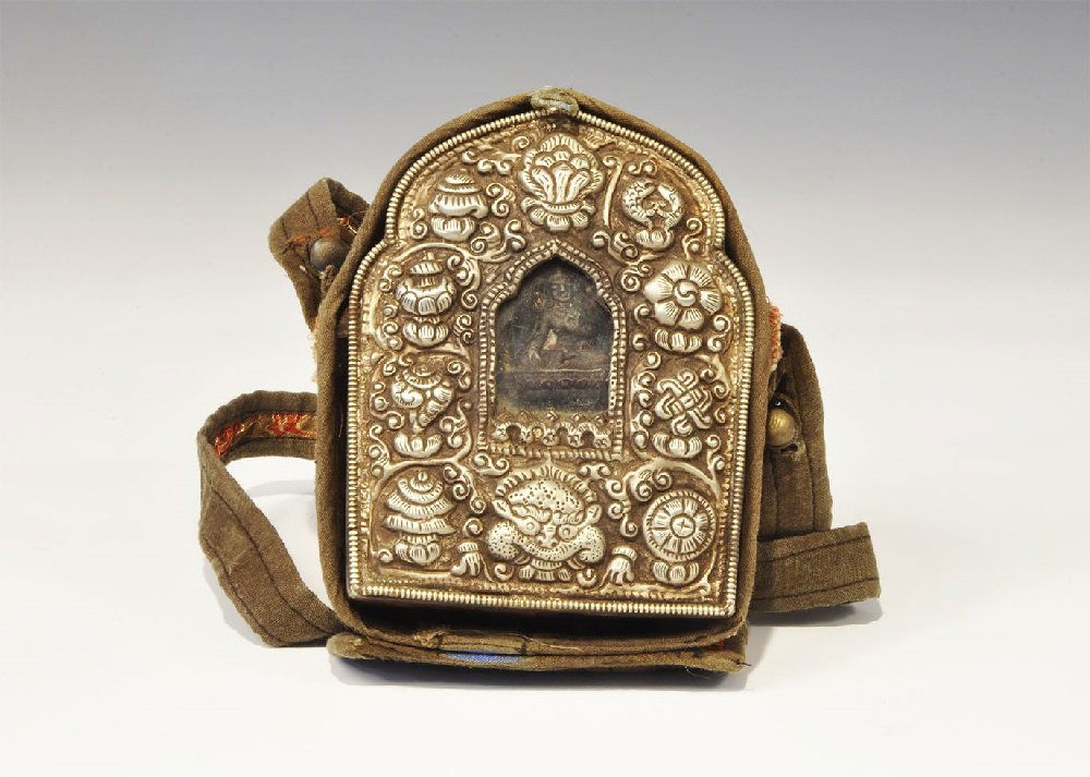 Chinese Tibetan Style White Metal Reliquary Box with: An undated archaistic repousse white metal box with glass window and cast figure behind on fabric backing, loops to the sides; contained in a fabric satchel with strap, quilted lid and button closure.