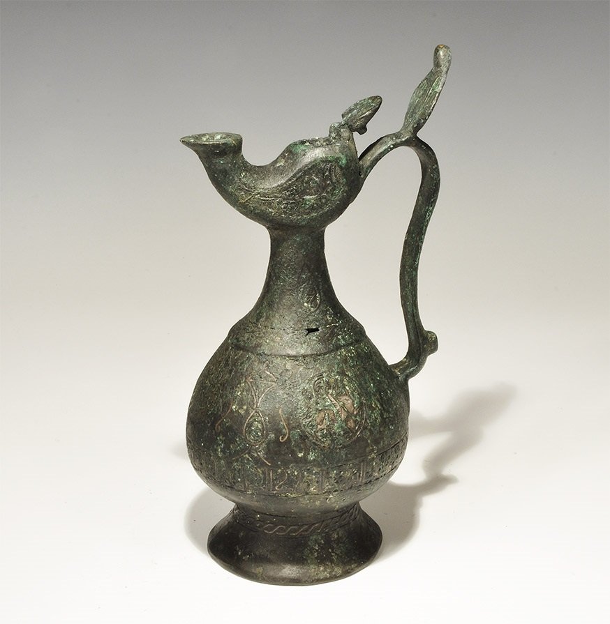 Islamic Style Bronze Calligraphic Ewer with Silver: An undated archaistic cast ewer with piriform body, flared base, curved strap handle with reflector, hinged lid and hexagonal-section spout; silver-inlaid calligraphic text to the body. 1.3 kg, 26 cm 