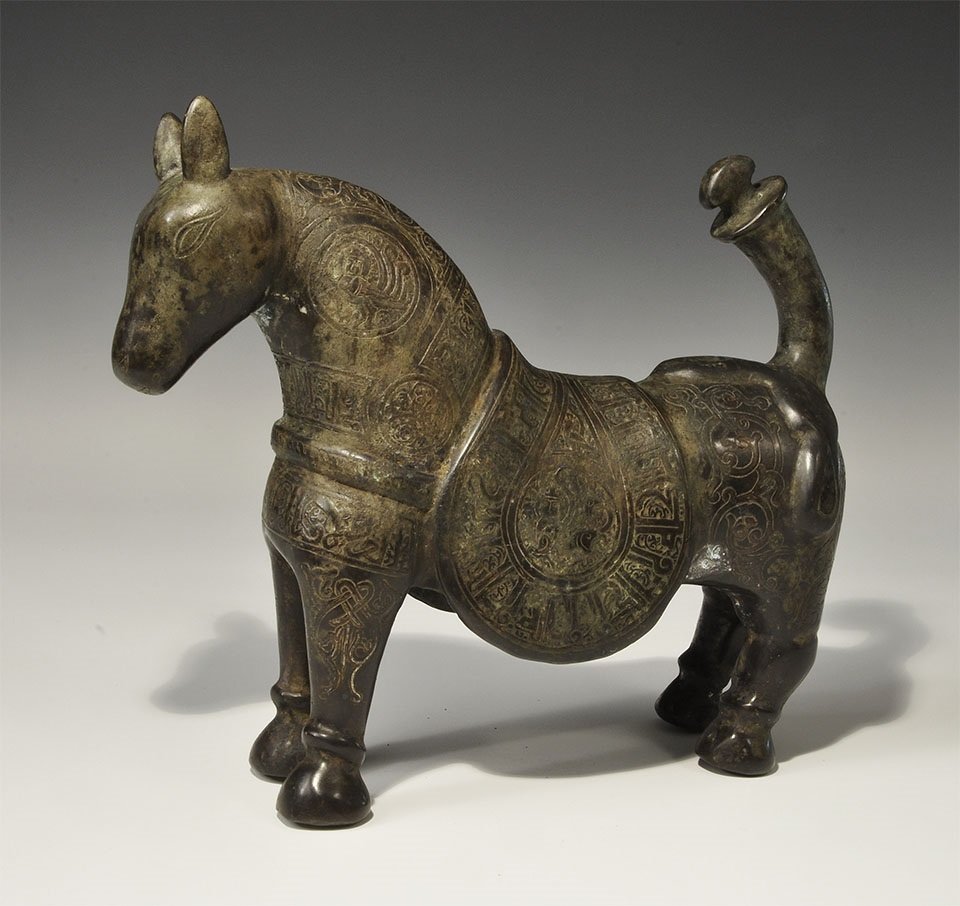 Islamic Style Bronze Horse with Calligraphic Silver: An undated archaistic cast horse figurine with saddle-cloth and chest-band; the surface ornamented with bands of silver-inlaid calligraphic text and scrolled foliage; the tail raised with discoid flan