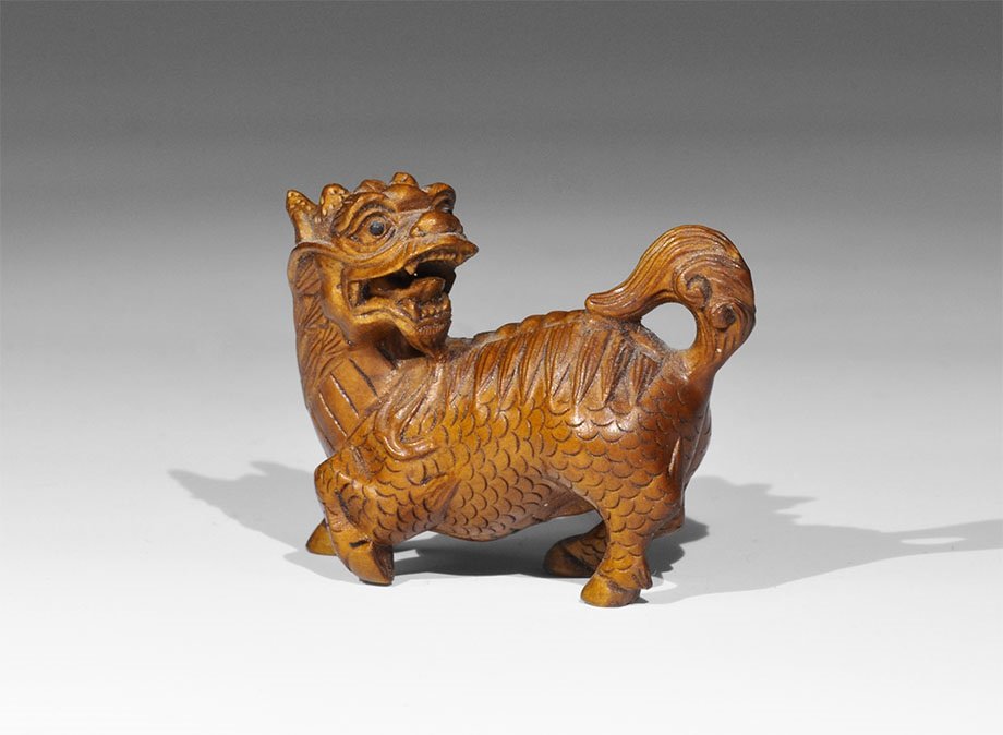 Japanese Antique Boxwood Kirin Dragon Netsuke: 20th century AD or earlier. A carved netsuke figurine of a regardant dragon with one raised leg, scrolled tail and open mouth; inset glass eyes; two holes to the underside for insertion of the cord; c