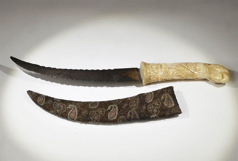 Indian Bone-Handled Dagger: An undated archaistic dagger with curved blade and zoomorphic handle; the blade with notched edges and gilt motif, unsharpened; the handle ornamented with bird-head finial; supplied with a fabric-cove