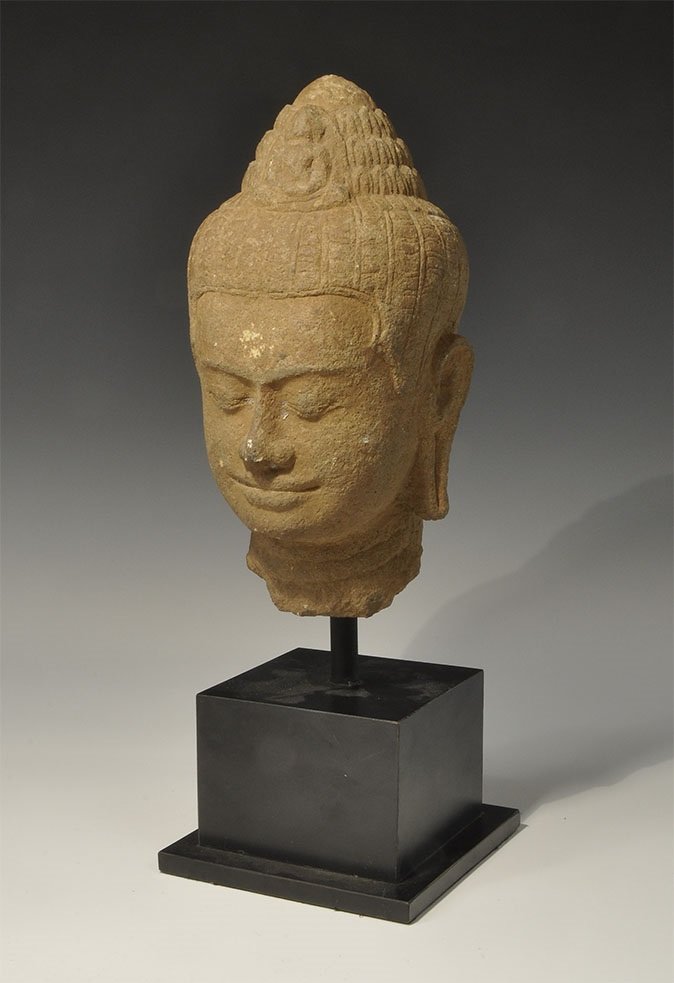 Cambodian Style Stone Buddha Head: An undated archaistic carved crowned head of the Buddha with serene smile and elongated earlobes; mounted on a custom-made stand. 4.9 kg, 36 cm including stand (14"). From a South East London collecti