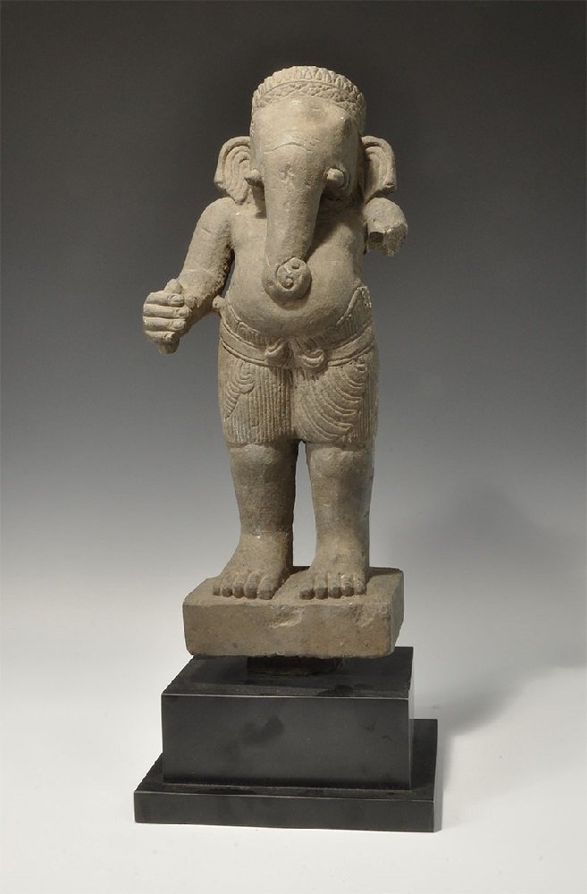 Cambodian Style Stone Ganesh Statuette: An undated archaistic carved statuette of Ganesh standing on a rectangular base, wearing a kilt, belt and conical crown; mounted on a custom-made stand. 7.2 kg, 47 cm (18 1/2"). Ex South East London c