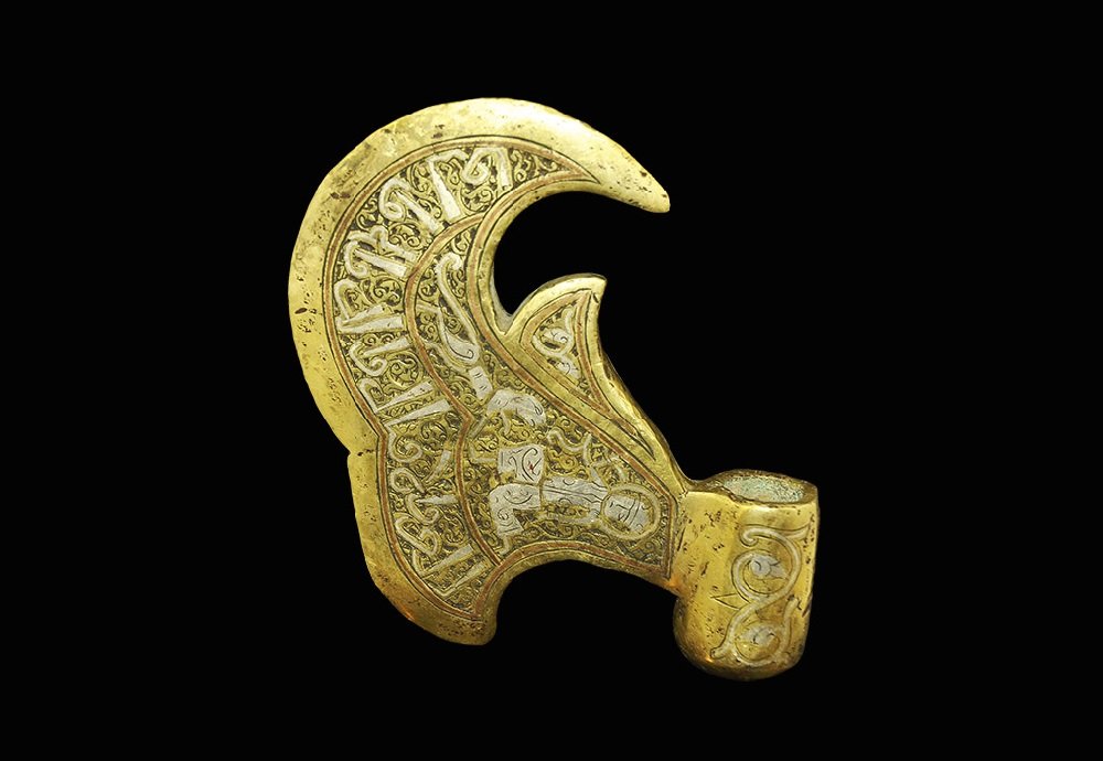 Islamic Style Bronze Silver-Inlaid Axehead: An undated archaistic cast axehead with barrel-shaped socket and hooked blade; silver-inlaid scroll to the socket, huntsman and dog to the blade and band of calligraphic text to the edge. 505 grams, 1