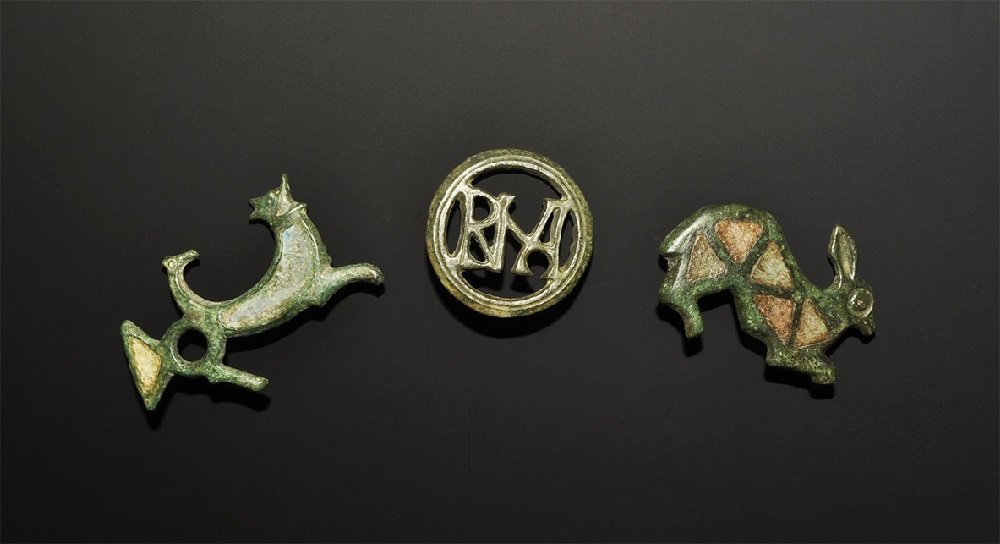 Roman Style Bronze Brooch Group: A mixed group of undated archaistic plate brooches comprising: a hare with triangular enamel cells; a cat and bird with enamel cells; an openwork disc with 'RNA' ligature to the centre. 47 grams total