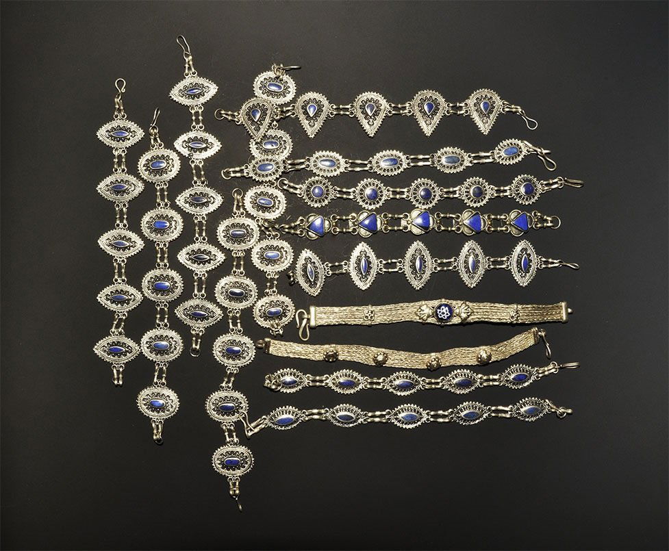 Indian Style White Metal Bracelet Group: A mixed group of undated archaistic chain bracelets, twelve with faux lapis lazuli inserts, one with flat-section strap and blue glass cabochon; one similar with white metal rosettes. 340 grams total,