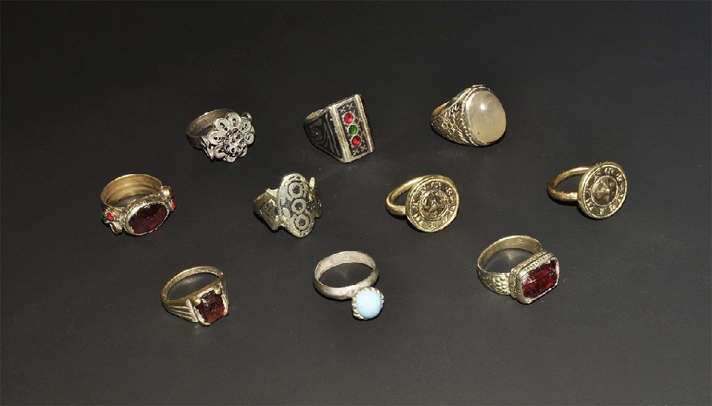 Post Medieval Bronze Finger Ring Group: A mixed group of undated archaistic rings comprising: one with rectangular bezel, red and green inserts; one with filigree rosette; two with faceted red glass inserts; one with elliptical bezel, geome