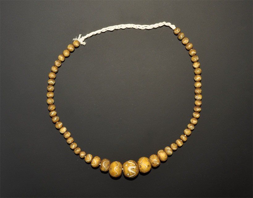 Near Eastern Gabari Style Glass Bead String: A graduated string of undated archaistic barrel-shaped beads in yellow with white marvered trails. 63 grams, 40 cm (15 3/4"). Property of a London collector, acquired early 1990s. [No Reserve]