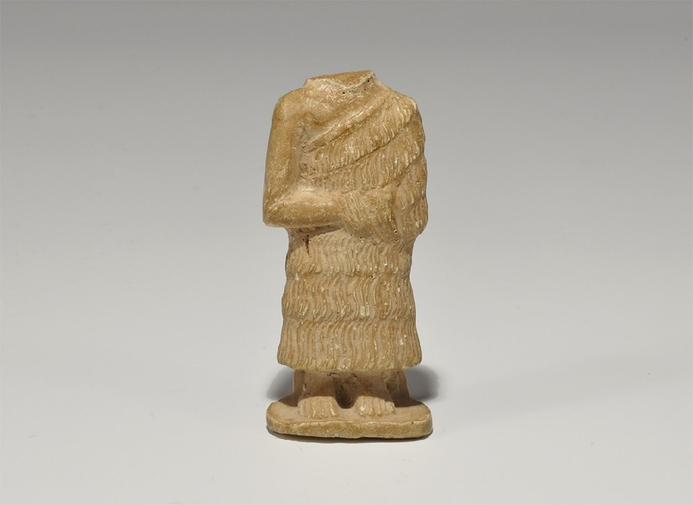 Near Eastern Syrian Style Stone Supplicant Figurine: An undated archaistic carved stone figurine depicting a standing figure in a shaggy cloak, the hands folded across the midriff, lower part of the hair to the reverse. 27 grams, 59 mm (2 3/4"). Propert