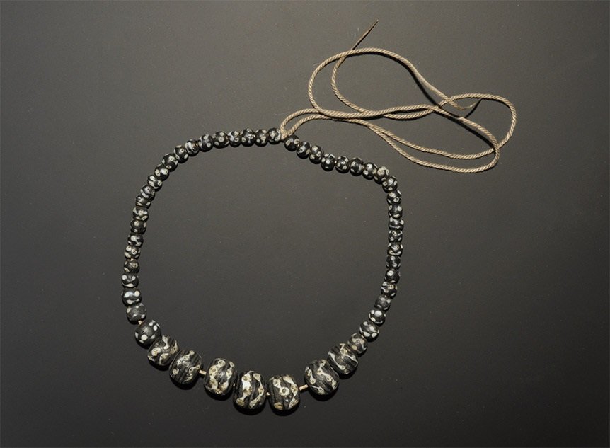 Near Eastern Gabari Style Glass Bead String: A graduated string of undated archaistic globular beads in black glass with marvered white trails. 58 grams, 36 cm (14 1/4"). Acquired on the London art market before 2000. [No Reserve]