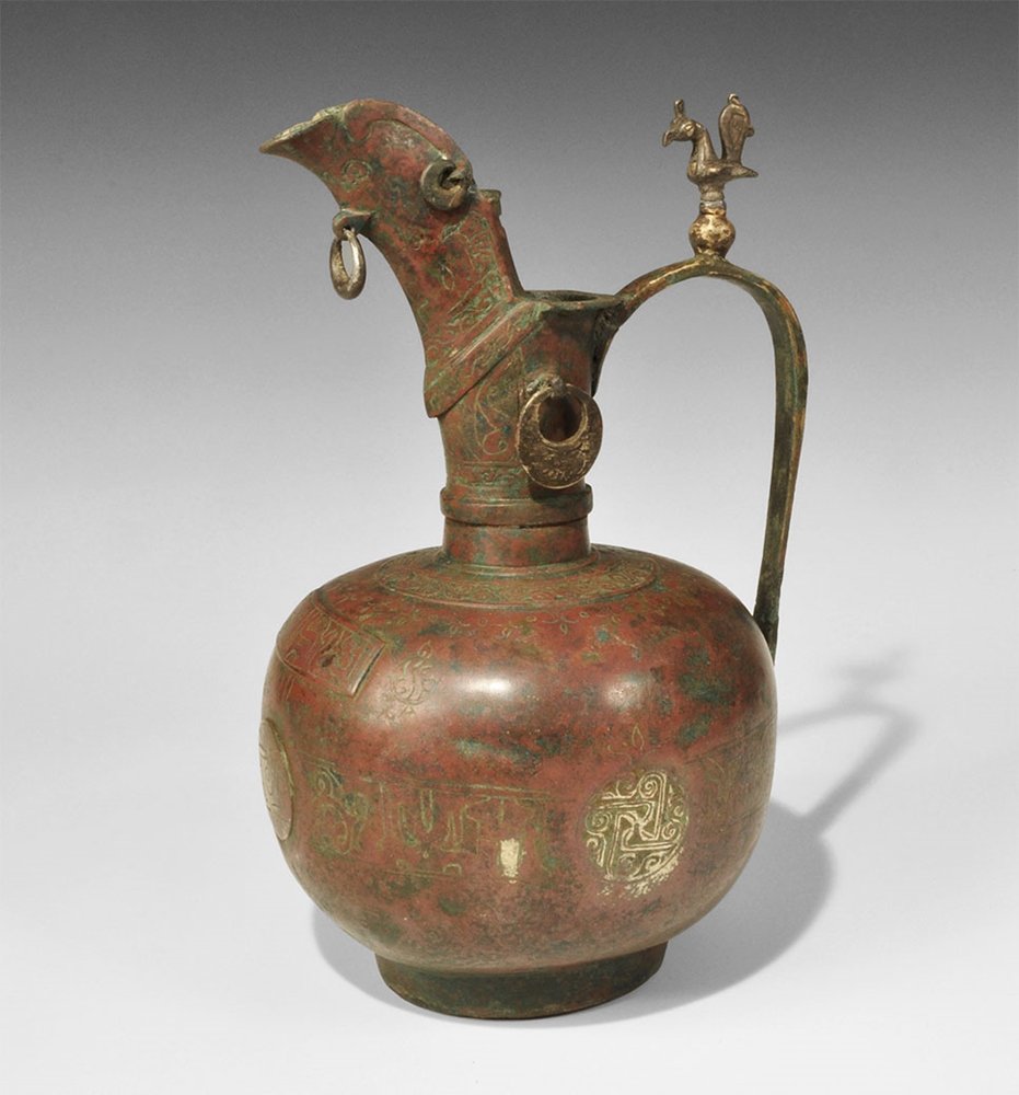 Islamic Bronze Calligraphic Ewer: 12th century AD. A cast globular ewer with discoid base, tubular neck with collar and everted rim, high open-back spout with grill; the strap handle with thumb-pad of a small model bird on a globe mod