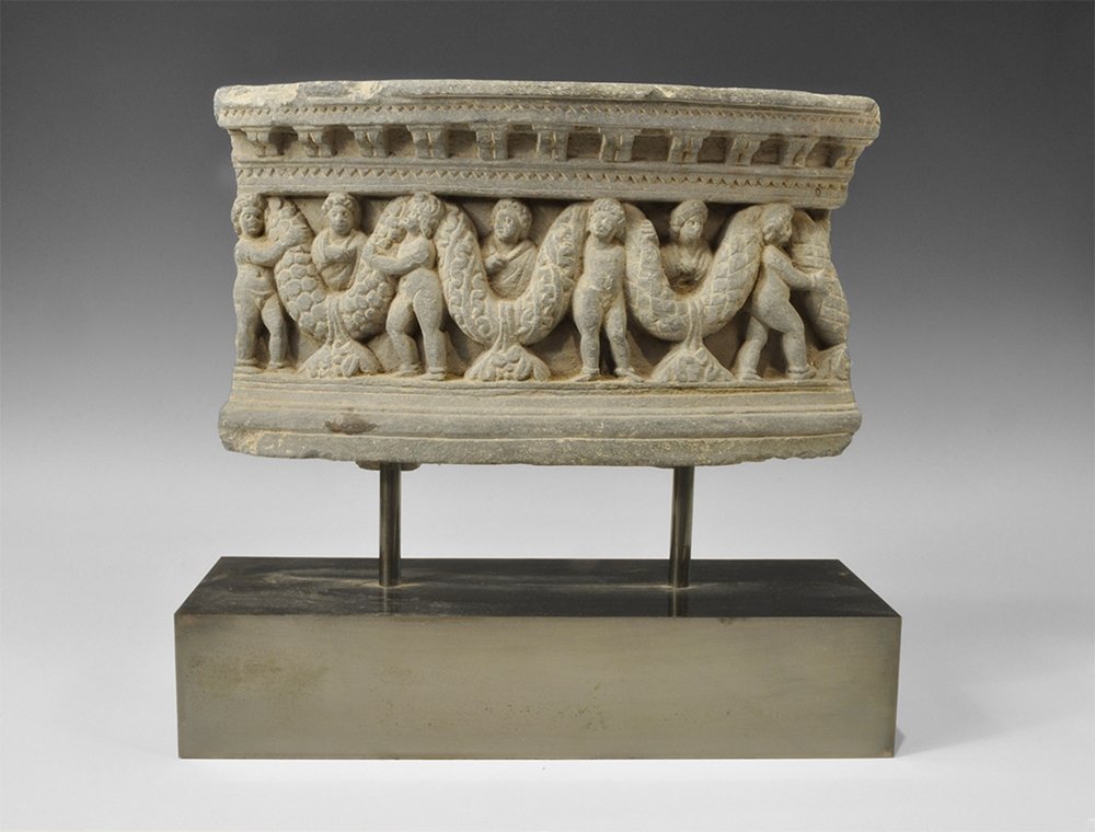 Gandharan Garland-Bearers Frieze: 2nd-3rd century AD. A carved schist porticoed scene of four nude garland-bearers with male busts between; slightly curved in plan, two incised characters to the upper face; mounted on a custom-made st