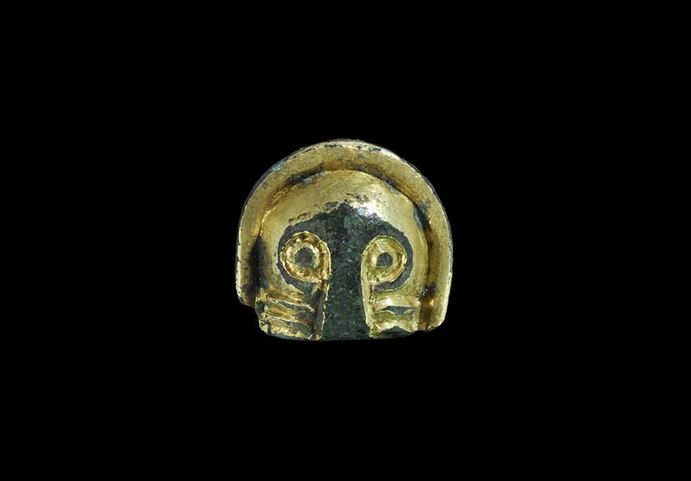 Anglo-Saxon Gilt-Bronze Chip-Carved Face Mount: 6th-7th century AD. A cast D-shaped mount of a human mask with pellet eyes, expanding bar nose and three lateral ribs to the lower face, hollow to the reverse; mounted on a custom-made stand. Cf. the