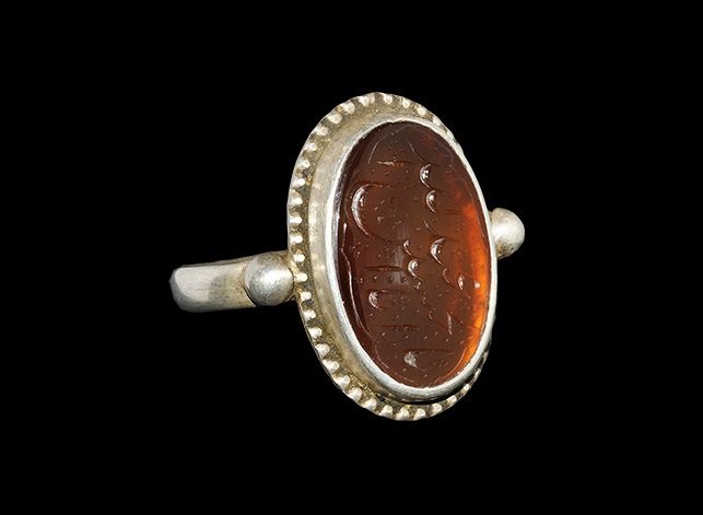 Islamic Stone Intaglio in a Modern Silver Finger Ring: An undated archaistic elliptical red hardstone intaglio with calligraphic text set into the bezel of a round-section hoop with pellets to the shoulders. 4.69 grams, 24 mm overall, 20.03 mm internal di