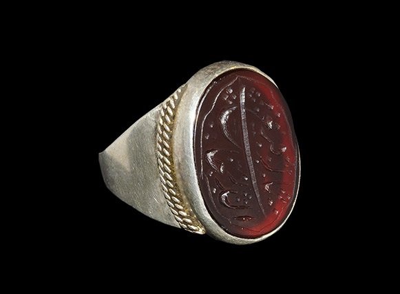 Islamic Stone Intaglio in Modern Silver Finger Ring: A flat-section hoop expanding to a discoid bezel with ropework trim at the junctions; red hardstone intaglio with calligraphic text. 6.47 grams, 24 mm overall,17.81 mm internal diameter (approximate s