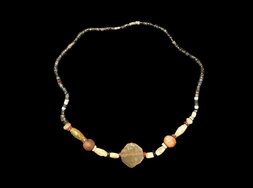 Near Eastern Mespotamian Stone Bead Necklace: 2nd millennium BC. A restrung necklace of small steatite annular beads with larger carnelian and other beads, central plano-convex crystal plaque. 71 grams, 64 cm (25 1/4"). Ex Steinberg collection; a