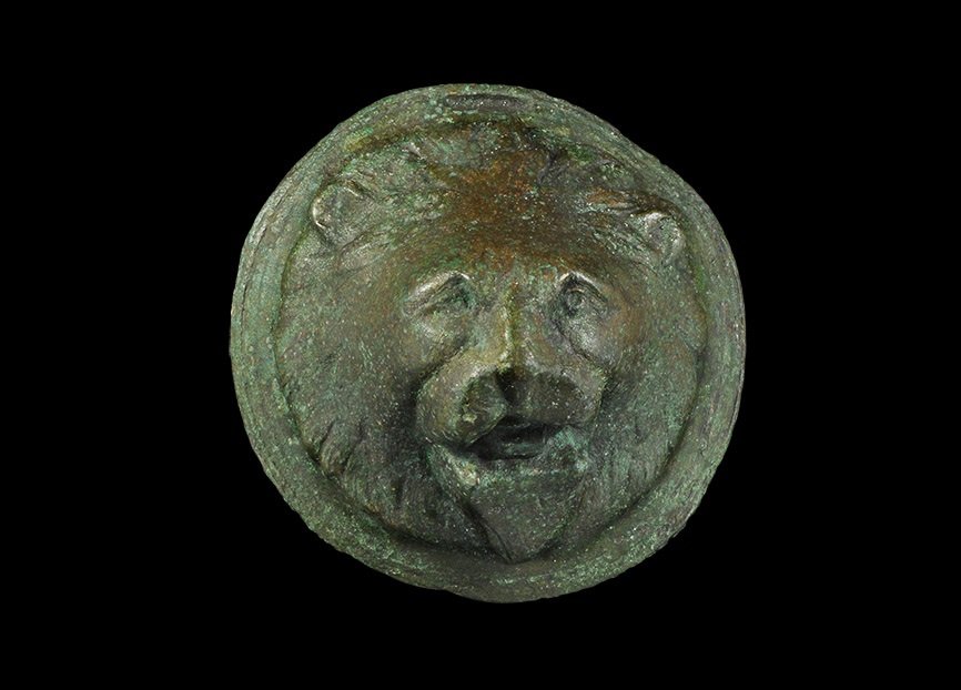Roman Bronze Lion-Head Mount: 1st-2nd century AD. A substantial cast mount formed as a lion's head with open mouth and radiating mane, flared flange rim; to the reverse, an iron mounting spike with angled tip. 263 grams, 88 mm (3 