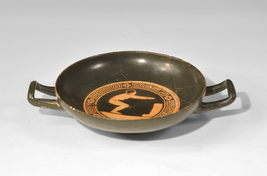 Greek Corinthian Style Painted Ceramic Kylix with: 19th century AD. A Grand Tour blackware kylix in 5th-3rd century AD style, with low base and two square handles; to the centre of the inner face a band of geometric ornament enclosing a crouching gymn