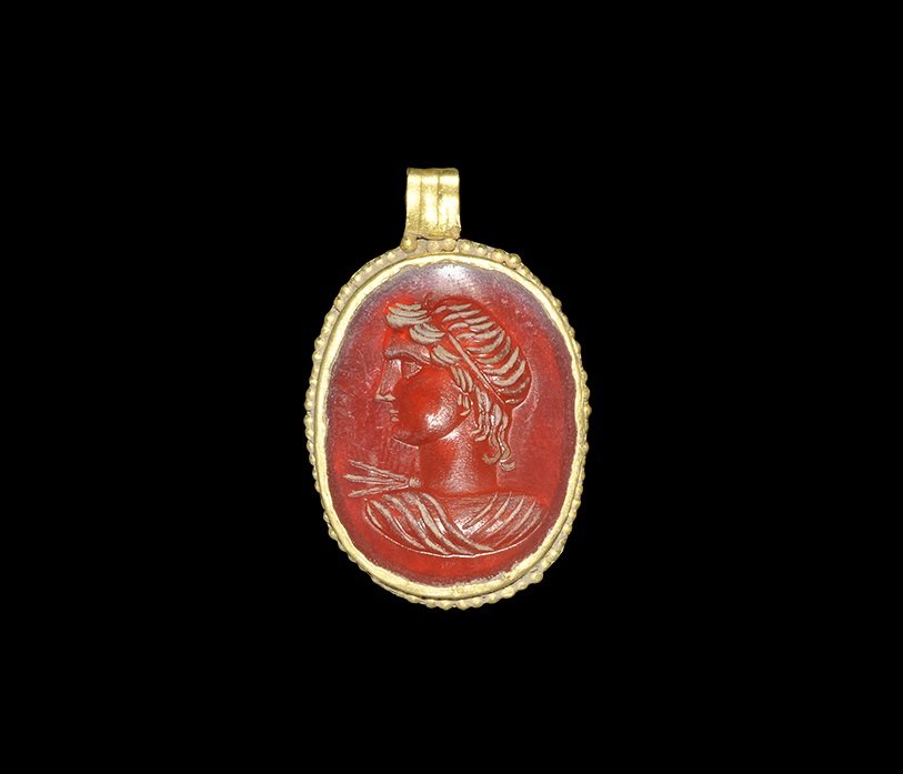 Roman Gold Pendant with Intaglio Bust of Apollo: 1st-2nd century AD. A discoid plaque in gold cell with granulation to the outer edge, ribbed suspension loop above; the plaque with intaglio of a youthful male profile bust, tunic to the upper body an