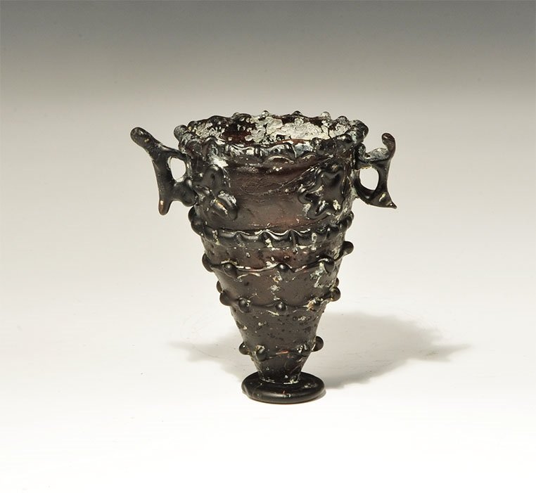 Islamic Style Black Glass Two-Handled Cup: An undated archaistic conical cup with applied discoid base, applied bands of ropework and lugs to the body and rim, applied rosettes below the rim, two lateral zoomorphic handles. 60 grams, 80mm (3 1