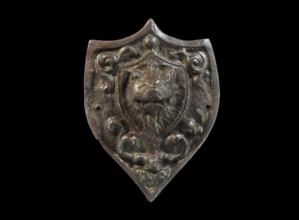 Post Medieval Bronze Coach Plate: 18th-19th century AD A cast heater-shield plaque with scooped upper edge, band of scrolled tendrils surrounding an escutcheon with lion's mask modelled in the half-round; pierced twice for attachment.