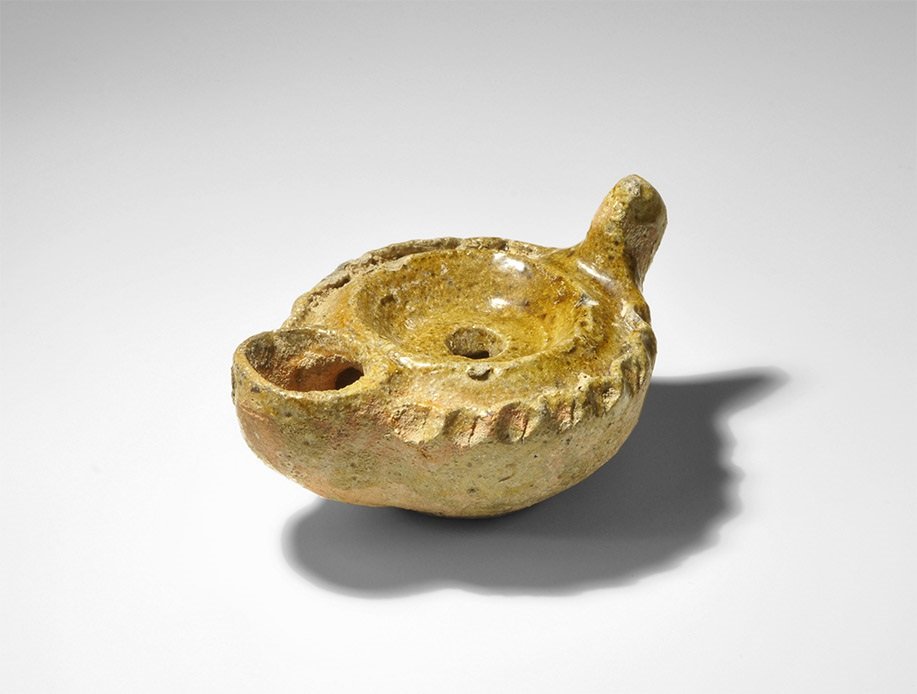 Islamic Ceramic Glazed Oil Lamp: 7th-8th century AD. A squat greyware oil lamp with piecrust shoulder, circular nozzle and knop handle, olive glaze to the upper face and sides. 122 grams, 98mm (3 3/4"). Property of a Hertfordshire, U
