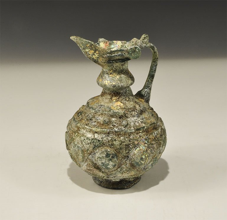 Islamic Style Glass Jug: An undated archaistic iridescent glass jug in 12th century style with low base, globular body, balustered neck, flared neck with spout; round-section handle to the rear, bands of disc and chevron orna