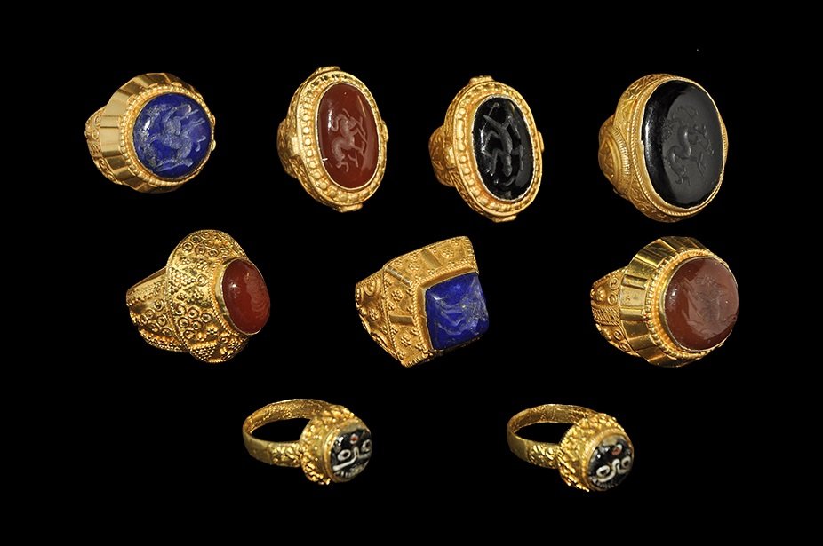 Near Eastern Style Gilt Ring Group: A mixed group of undated archaistic gilt metal rings, each with an inset cabochon intaglio, comprising: one calligraphic text; two mosaic glass faces; three dragons; three quadrupeds. 182 grams total,