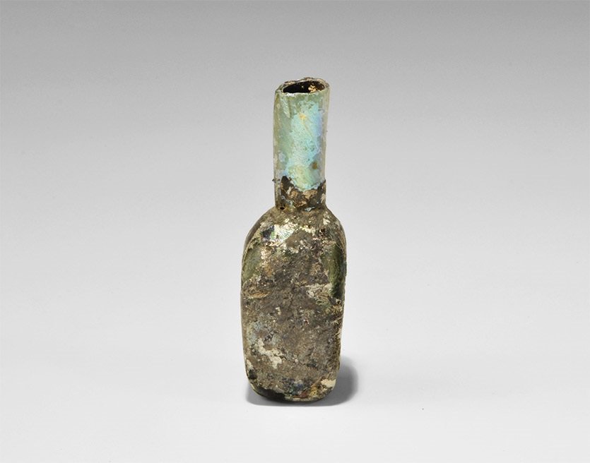 Roman Iridescent Glass Bottle: 2nd-3rd century AD. A mould-blown miniature bottle, square in plan with tubular neck, and cut rim. 28 grams, 72 mm (2 3/4"). Property of a retired sculptor; from a collection formed in the 1980s.