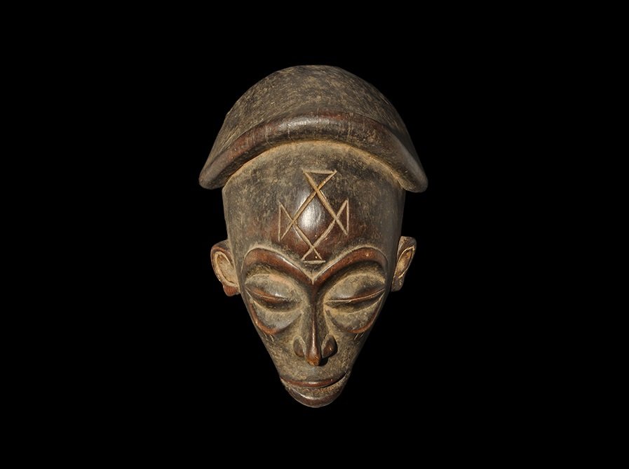 Ethnographic West African Wooden Carved Mask: Chokwe tribe, 20th century AD. A carved mask with slit eyes, open mouth, headdress with raised rim, cruciform scarification to the brow; reverse hollowed, hole to accept a mounting pin. 311 grams, 28c