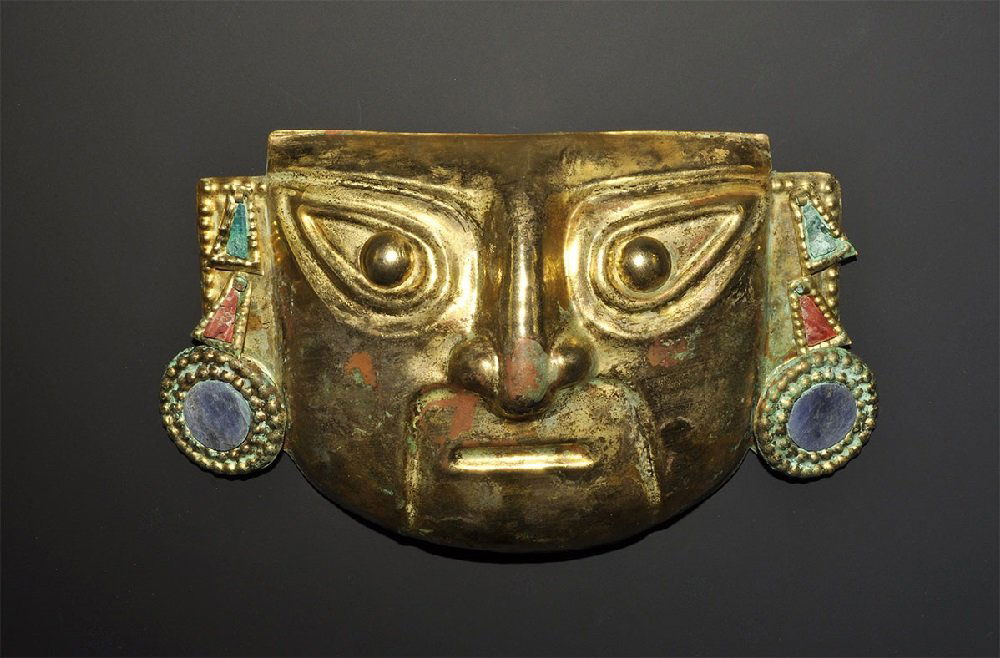 Pre-Columbian Moche Gilt-Bronze Mask with Inlays: 100-700 AD. A sheet gilt metal mask with raised detail to the eyes and nose; separate appliqués to each ear, two trapezoidal in red and green stone and a third a discoid blue stone, each on a gilt me