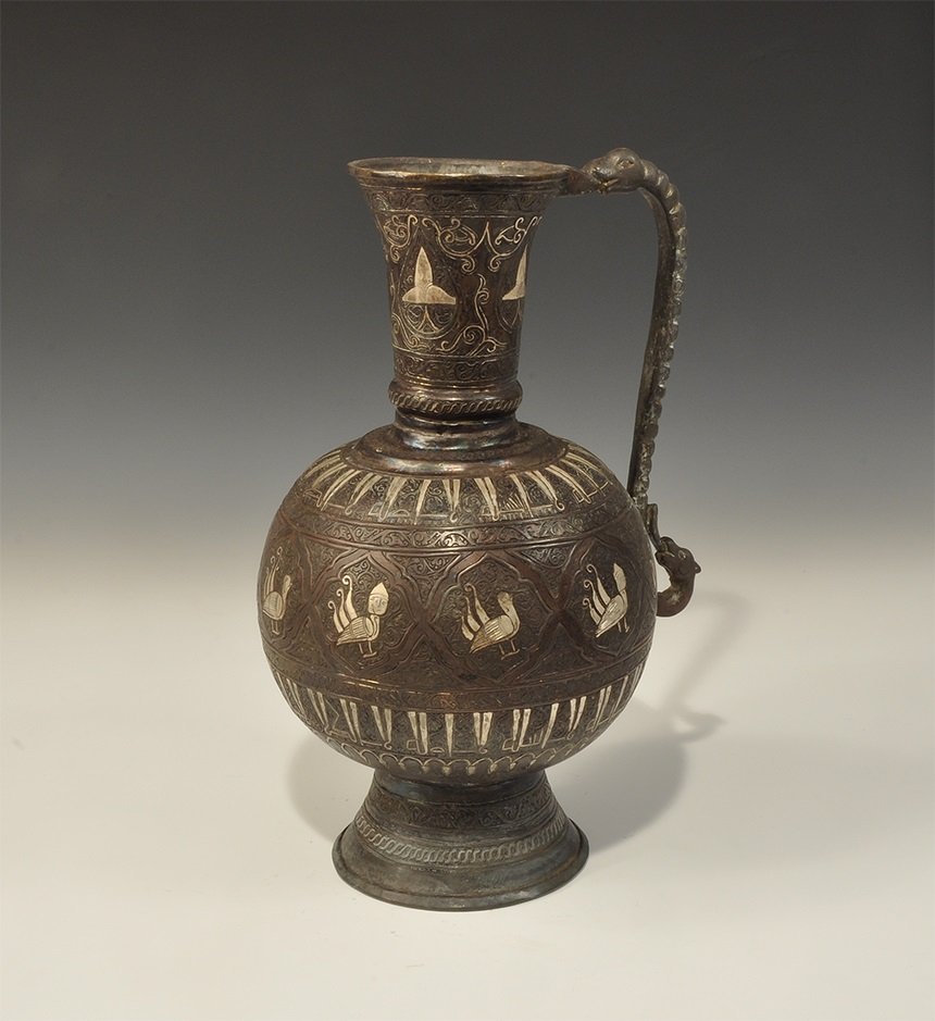 Islamic Bronze Jug with Calligraphic Silver Inlay: An undated archaistic Bidriware jug with globular body, domed foot, funicular neck and everted rim; scrolled zoomorphic handle to the rear; panels of inlaid silver trefoil and tendril ornament to the 